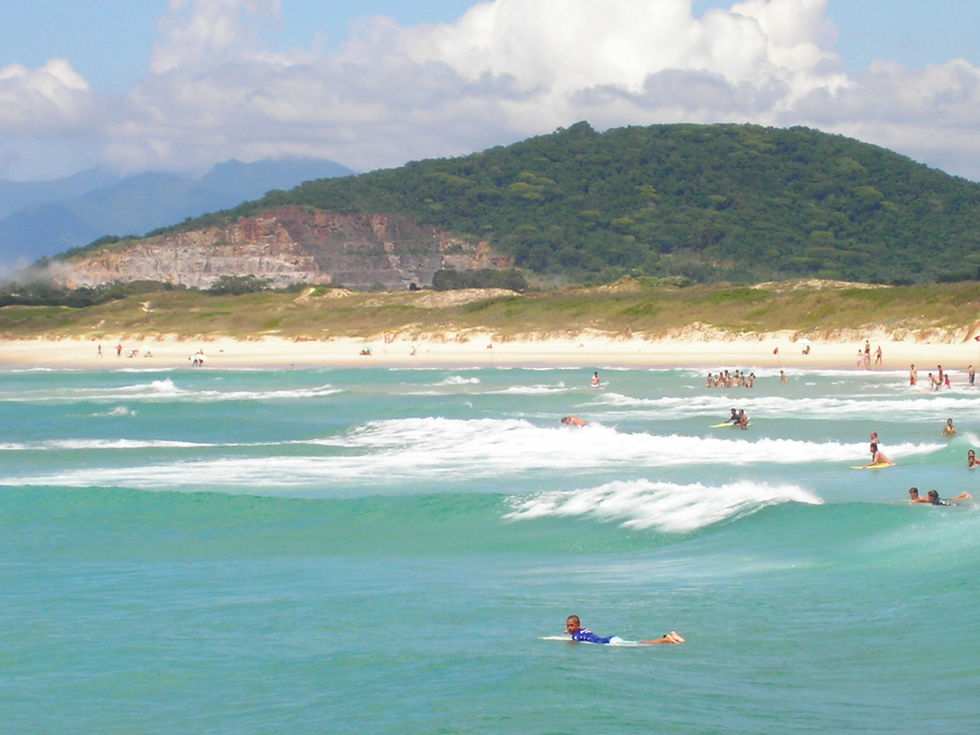 The waves play with the light, reflecting shades of emerald and turquoise. Upon them, surfers glide like birds on the wind, while in the background, the mountain rises with its green, rocky skin, guarding the secrets of time. It is a sea that invites, a horizon that enchants.