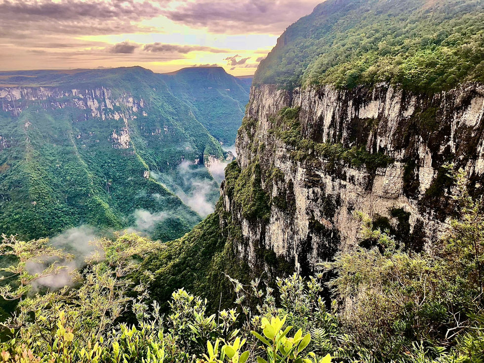 O sol se põe por trás das imponentes falésias, enquanto o verde da vegetação se entrelaça com o cinza das pedras. O ar fresco traz consigo a paz do vale, onde as nuvens se estendem suavemente pelo abismo.