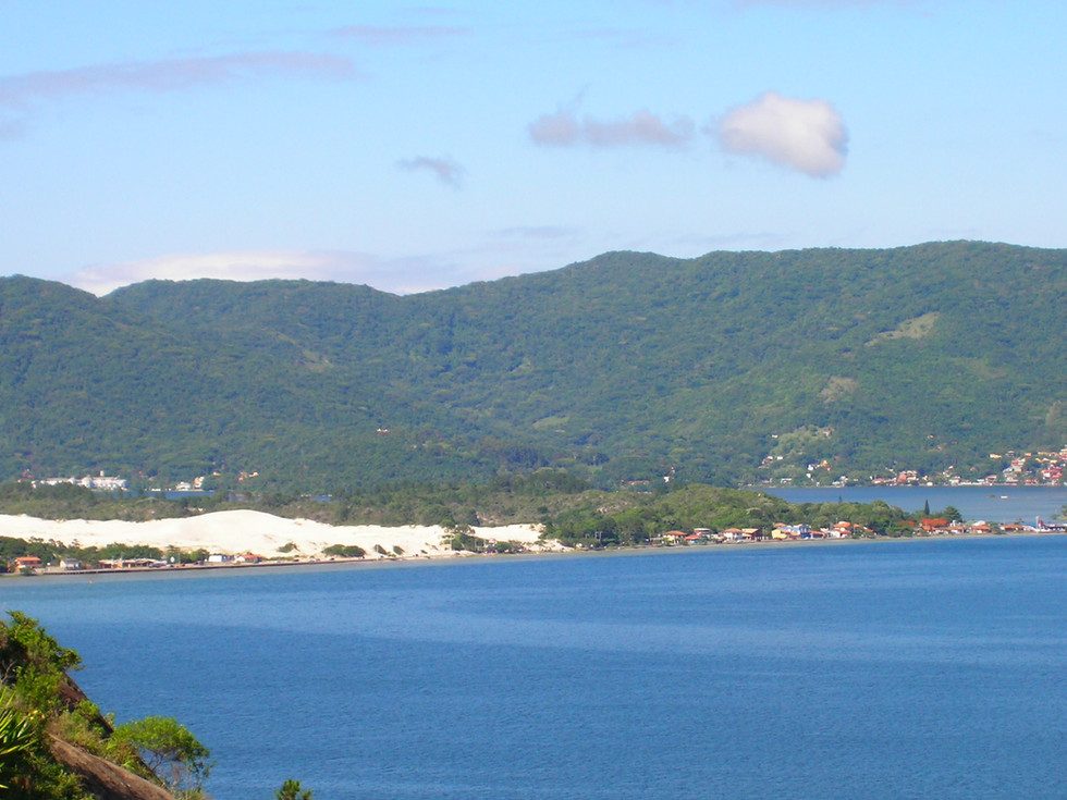 A imensidão azul abraça as dunas que brilham ao sol, como ouro em pó espalhado pela costa. Montanhas cobertas de verde assistem à cena, eternas, enquanto pequenas casas coloridas se agrupam na beira d’água, testemunhas discretas dessa poesia natural.