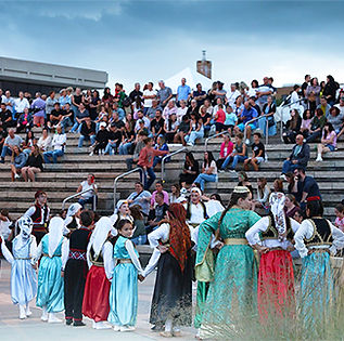 Crowd of people on Amphitheatre seating with a group of teens and children in traditional clothing preforming on stage. 
