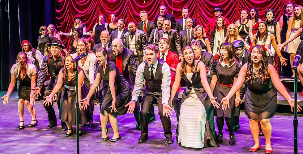 Image of a large group of singers performing on stage. Red swagged curtains hang behind them. Purple lights shine down and light the singers and the floor. Performers in the front are dancing. They are huddled in a bunch, knees bent, with hands outstretched. The singers are all wearing red, black, and white clothing, each in unique styles. All are singing, and most of them are smiling.