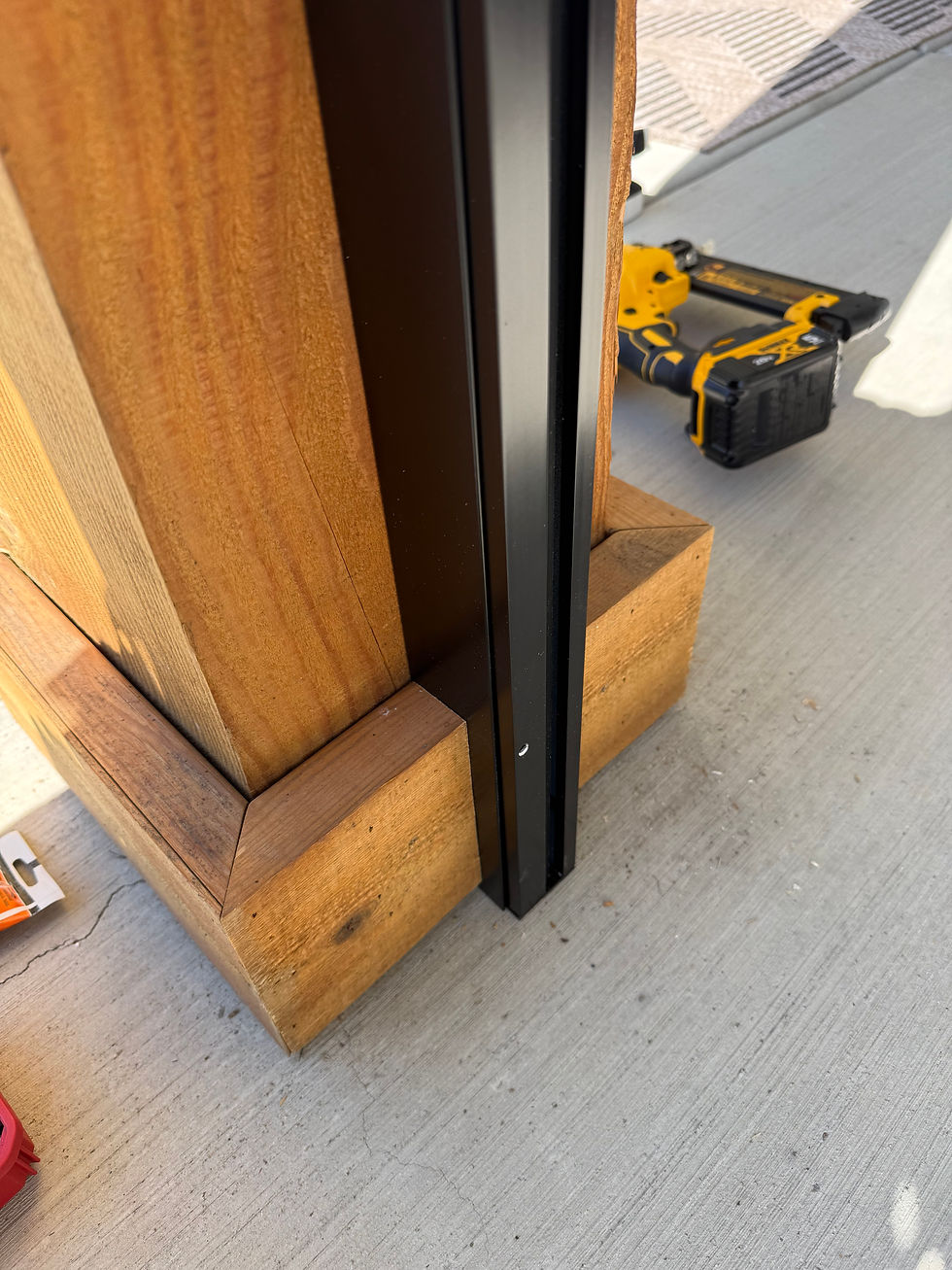 Close-up of a wooden post and black metal zipper track on concrete, with a yellow nail gun in the background during construction.
