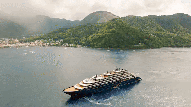 Cruise ship sailing near lush green mountains and a coastal town. The sky is cloudy, and the sea is calm, creating a serene atmosphere.