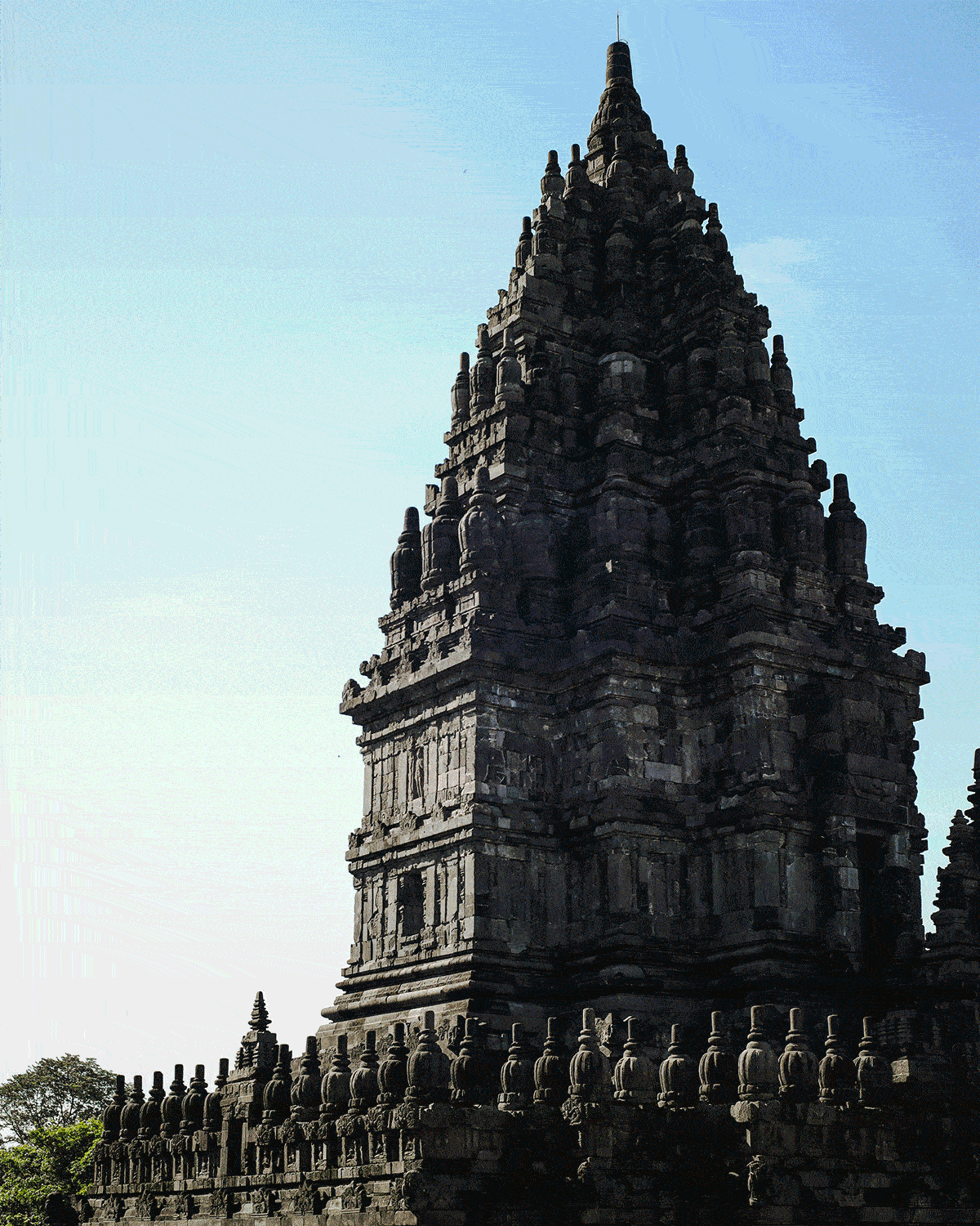 Ancient stone temple tower against a clear blue sky, with intricate carvings and a serene atmosphere, surrounded by lush trees.