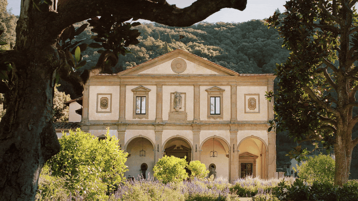 Historic building with arches and statues, set against a forested hill. Framed by trees, vibrant green bushes, and purple flowers.