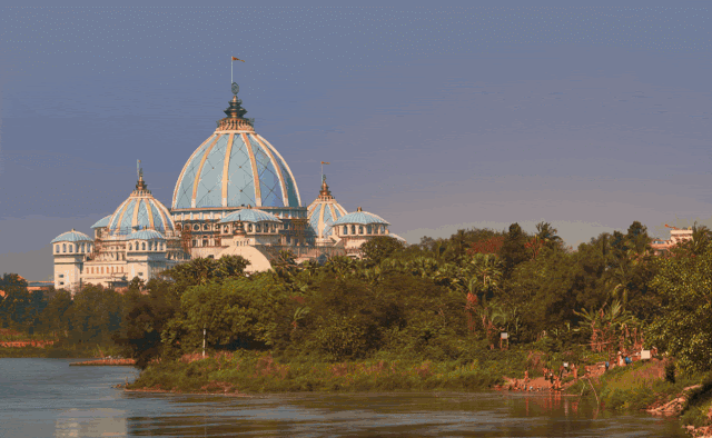 A grand temple with blue domes and tall spires near a calm river and lush greenery under a clear blue sky.