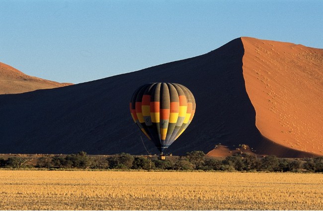 A colorful hot air balloon floats near large red dunes against a clear blue sky, with dry grasslands and scattered bushes in the foreground.