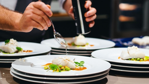 Chef garnishes plates with sauce in a kitchen. White plates hold fish, greens, and orange puree. Hands hold a spoon and shaker.