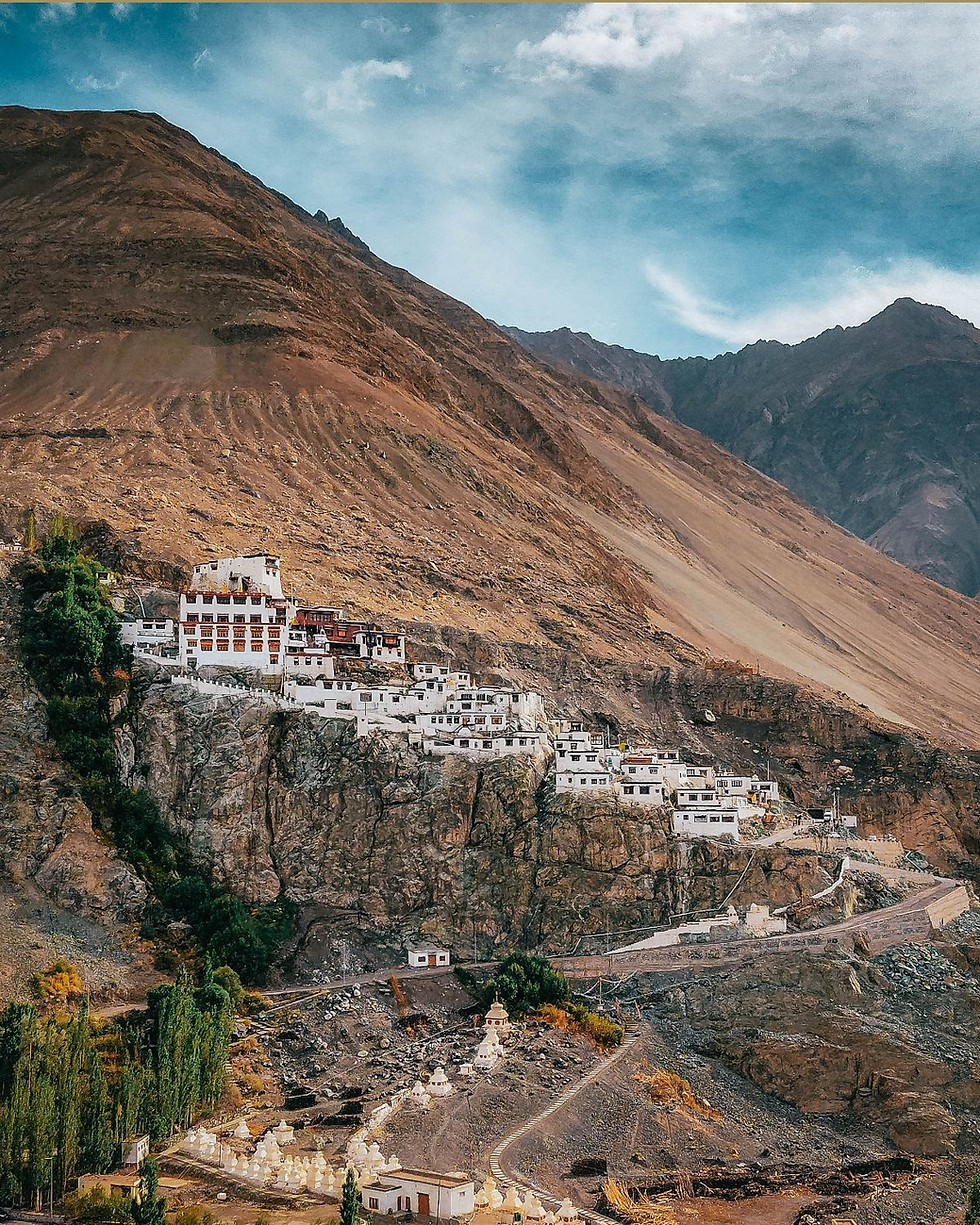 White monastery buildings cling to a rocky mountainside under a blue sky, surrounded by sparse greenery and rugged terrain.