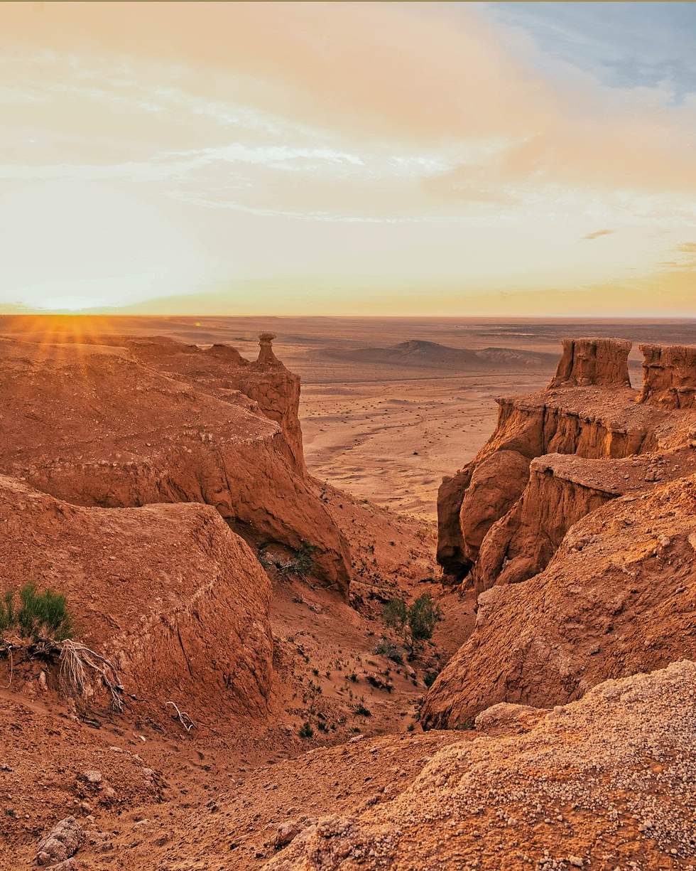Desert landscape at sunset with red rock formations, a wide valley, sparse vegetation, and a warm, glowing sky.