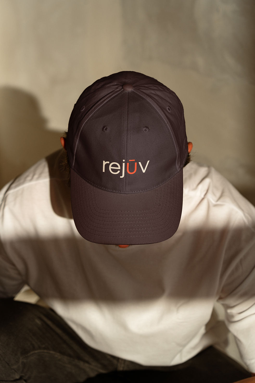 A person wearing a charcoal baseball cap with the Rejuv logo in cream, over a white top against a neutral wall.