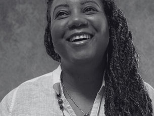 Smiling woman with braided hair in a white shirt, wearing a beaded necklace and heart pendant, against a textured gray background.