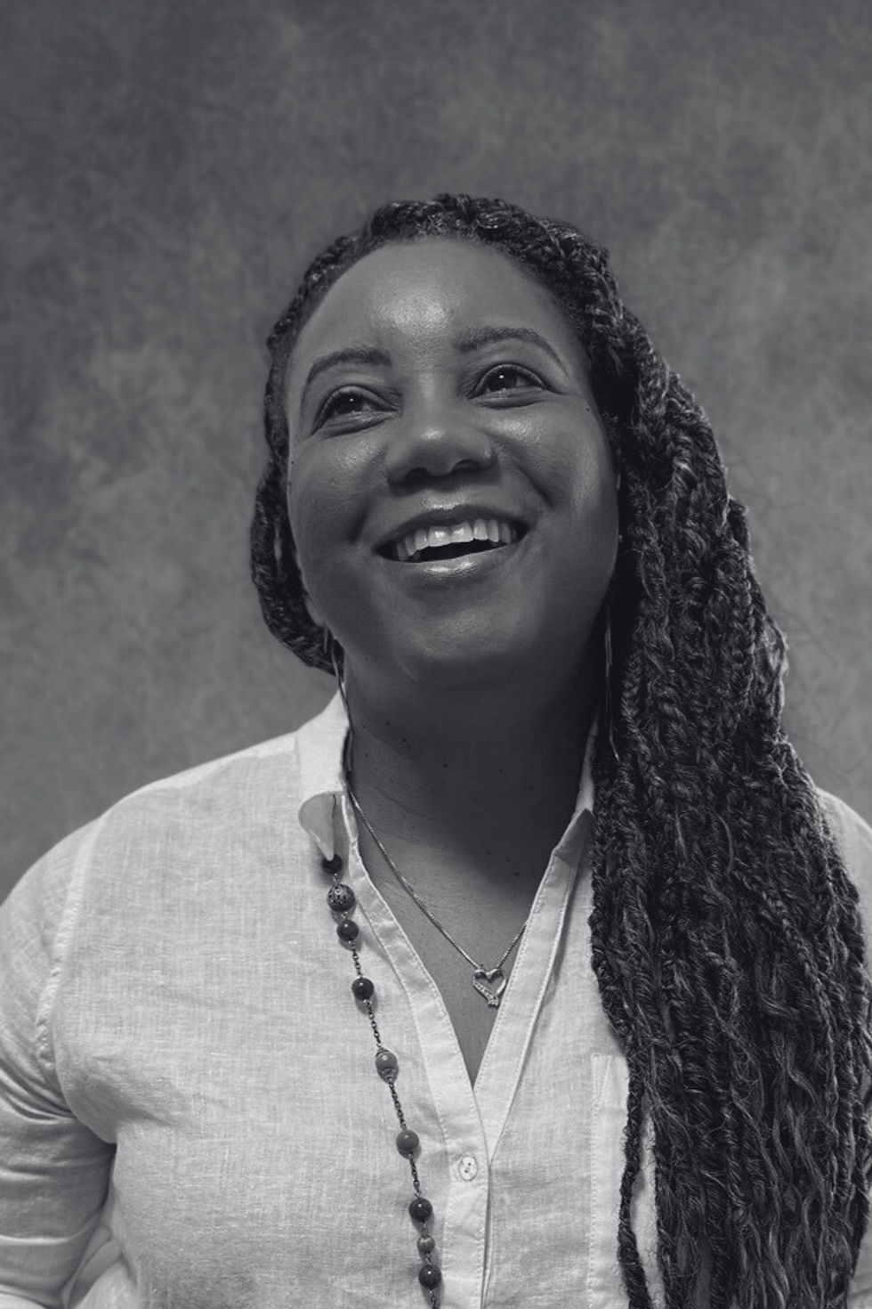 Smiling woman with braided hair in a white shirt, wearing a beaded necklace and heart pendant, against a textured gray background.