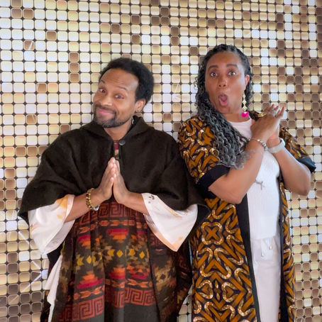 Two adults in patterned robes pose before a tiled wall, one smiling with palms together, the other wide-eyed with hands clasped.