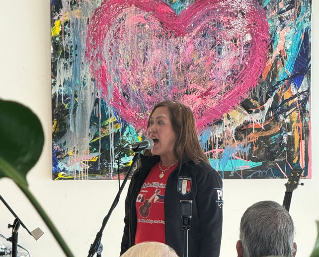 Jeanette Perez. A woman standing at a microphone and speaking to the audience. Abstract art is in the background.