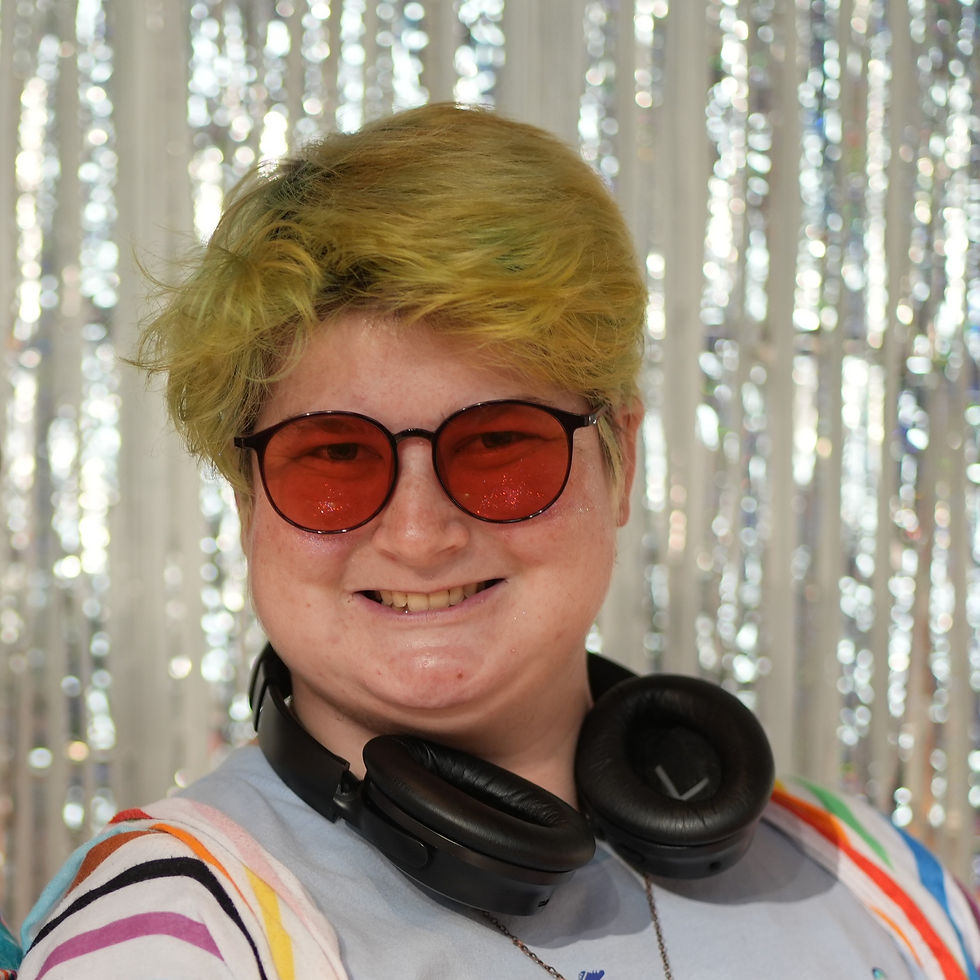 Remiel Cooper- a fat white person smiles at the camera. Remiel has short green hair that has faded past its prime. They wear red tinted sunglasses with black rims that make it hard to tell the color of their eyes. beneath their sunglasses you can see glitter glowing on their cheeks. Around their neck their is a pair of black noise cancelling headphones.