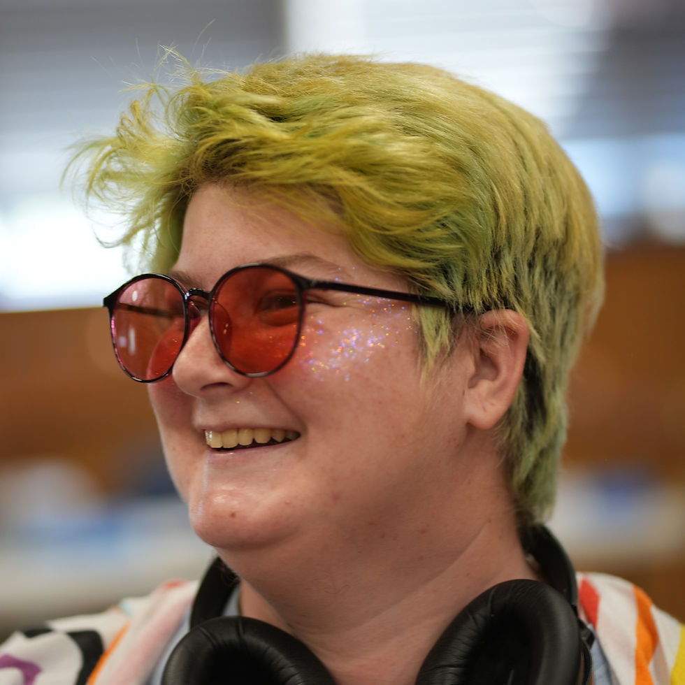 Remiel smiles widely looking oof camera. Their hair is a faded grin and their is body glitter on their cheeks. They wear red tinted sunglasses. Part of their nose cancelling headphones can be seen around their neck.