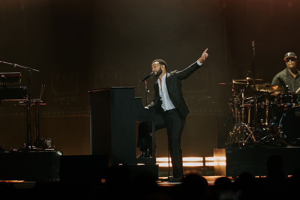 John Legend in a black suit sings passionately at a piano on stage, with a drummer in the background. Warm lighting creates a dramatic mood.