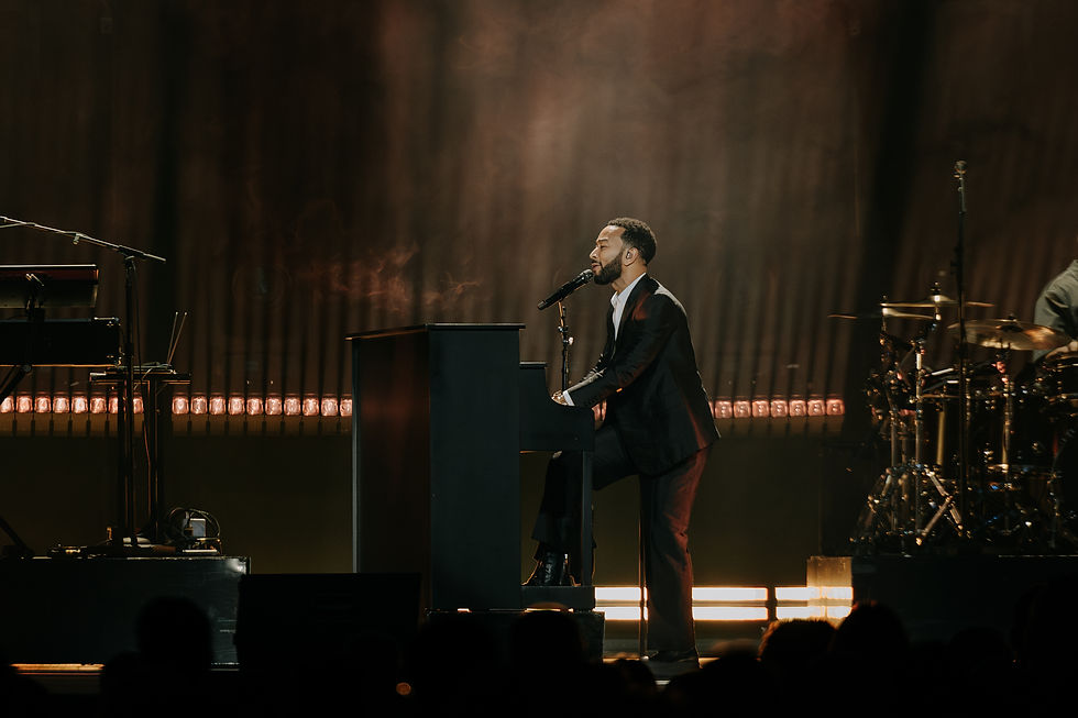 John Legend plays piano while singing onstage. Dimly lit with warm lights; drum set and microphone visible in background.