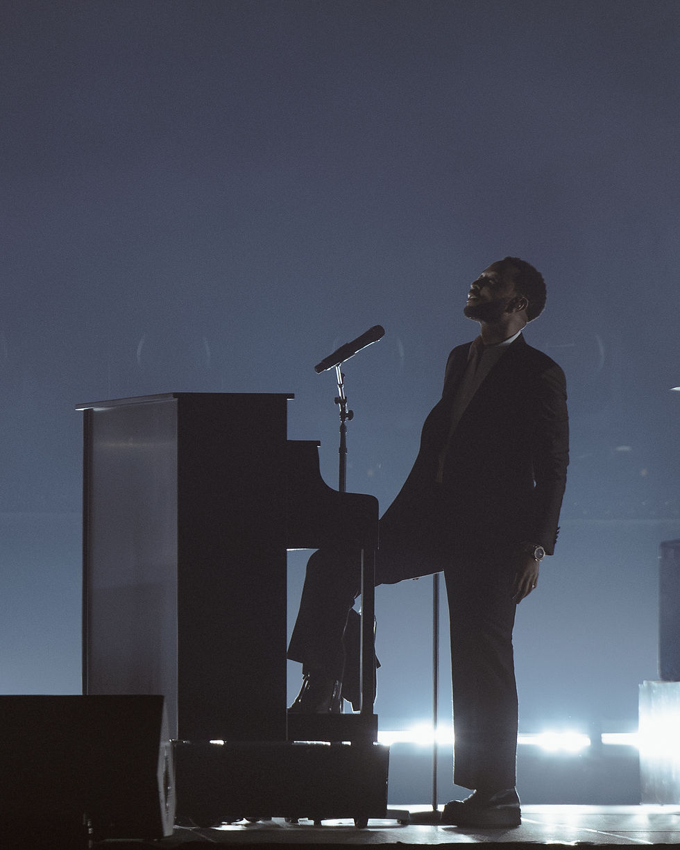 John Legend Silhouetted in a suit stands by a piano with a microphone, set against a blue-lit stage, exuding a contemplative mood.