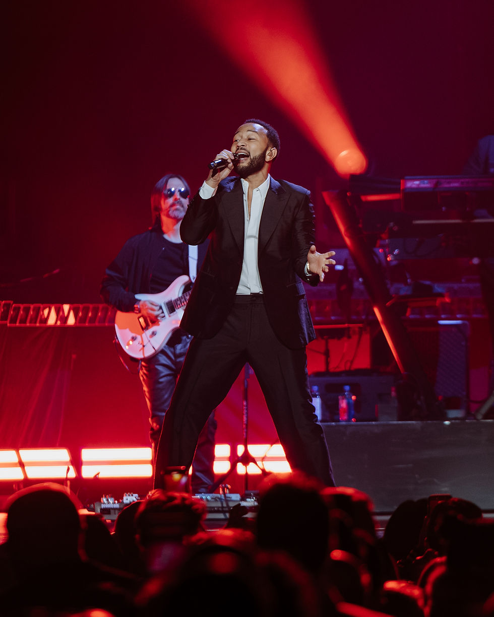 John Legend in a black suit passionately performing with a microphone on stage. Red lights illuminate the background, guitarist in sunglasses behind.
