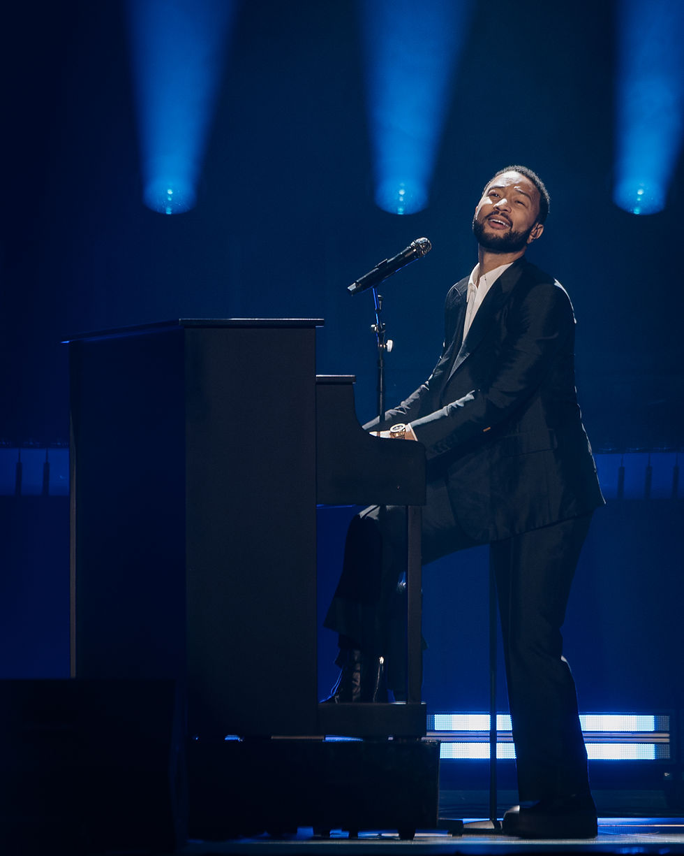 John Legend at Rogers Arena playing on a black piano under blue stage lights, expressing emotion while performing.