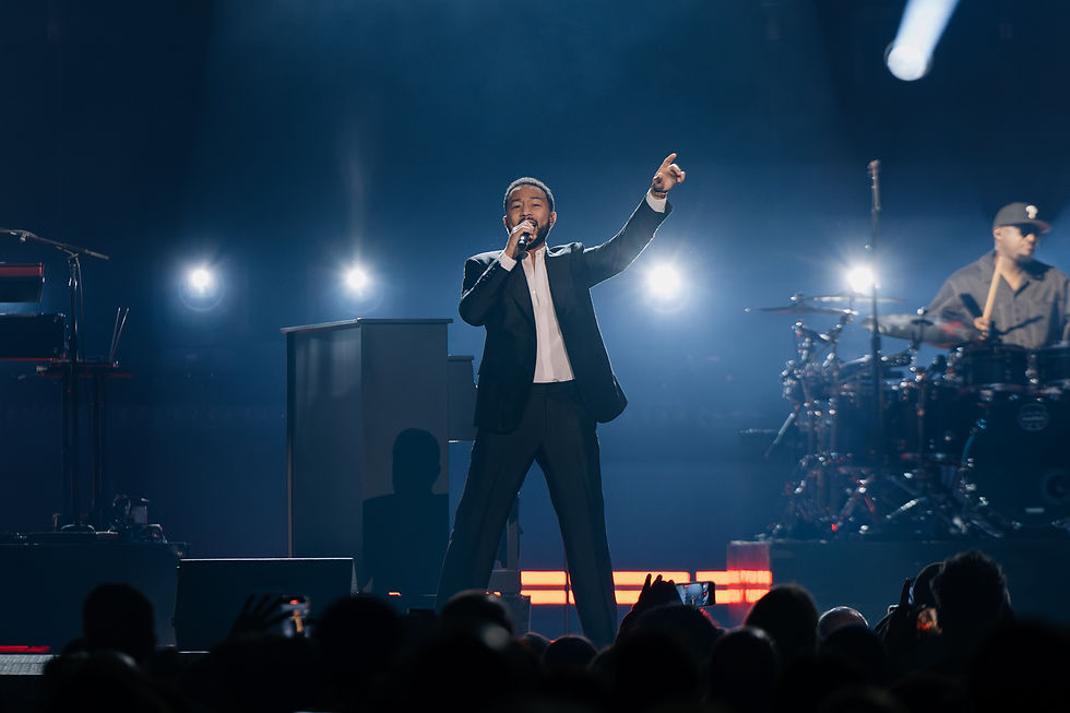 John Legend in a black suit performs on stage with a microphone. Drummer in the background, blue lighting, energetic atmosphere.