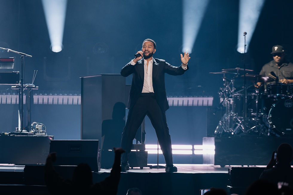 John Legend performs energetically on stage, holding a microphone, with a band visible. Bright stage lights create a dynamic atmosphere.
