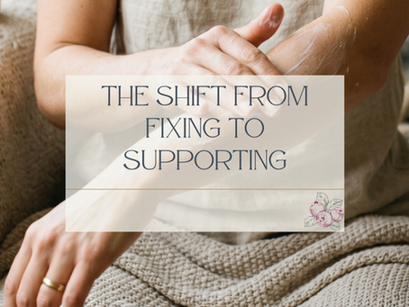 A woman in a natural linen tank top applies lotion to her forearm while seated on a textured neutral couch, a woven blanket across her lap. A gold ring is visible on her hand. The image is cropped to focus on her arms and torso. A semi-transparent cream overlay in the center displays the blog title "The Shift From Fixing to Supporting" in dark slate serif lettering, with a small Juneberry Apothecary botanical berry illustration in the lower right corner of the text box.