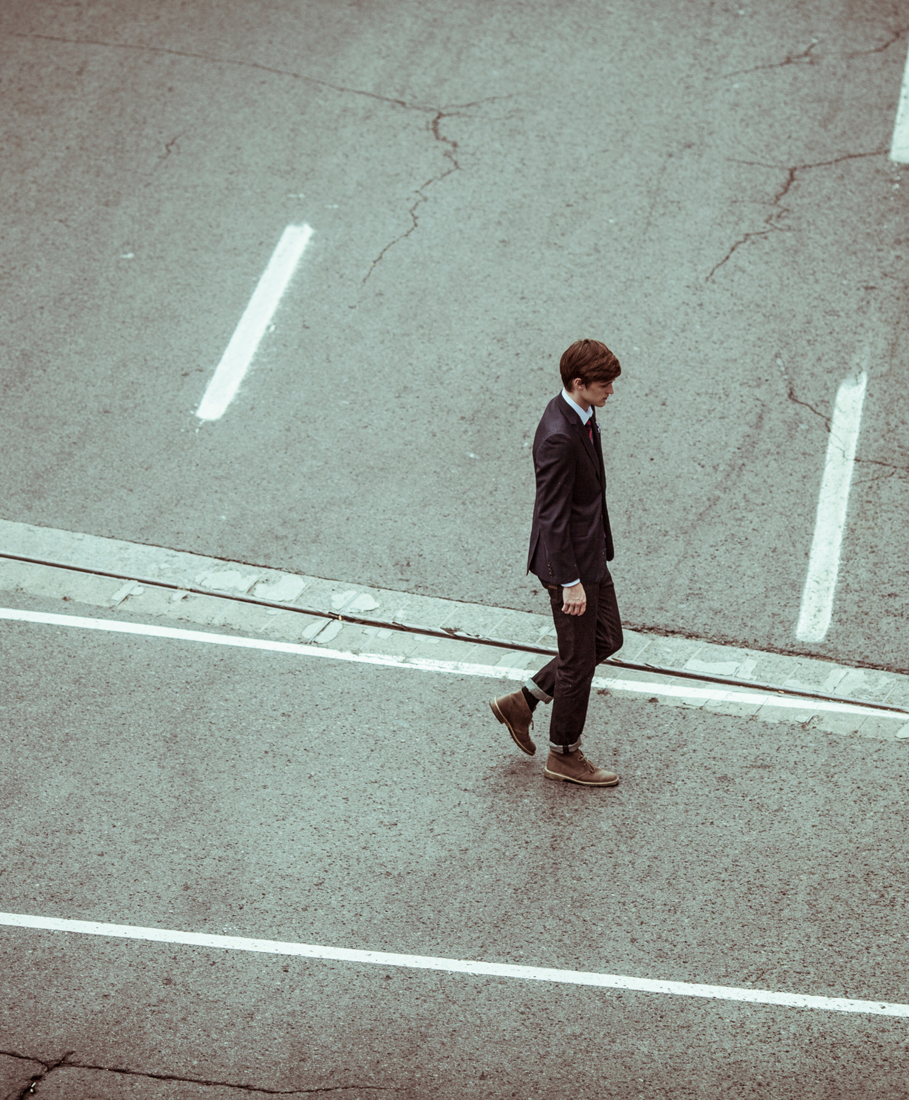 A suited man walks alone across a cracked empty road with white lane markings, seen from above in a quiet urban setting.