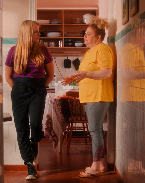 Frame horizontal de duas mulheres conversando na cozinha. Vistas emolduradas pela parede de um corredor de ladrilho, duas mulheres uma de frente para a outra conversando. A da esquerda está apoiada na parede, é loira, tem cabelos longos e lisos, usa camiseta roxa, calça preta e all star de cano alto preto. A da direita é gorda, tem cabelos cacheados loiros pretos no alto da cabeça, usa camiseta amarela, leggin cinza e chinelo. Ao fundo, uma mesa e um armário com utensílios domésticos.