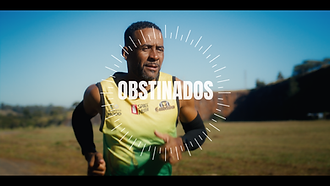 Frame horizontal de um homem negro praticando corrida. Em primeiro, visto do peito para cima um homem negro de cabelos castanhos curtos e cavanhaque, usa uma camiseta regata com degradê  do amarelo para o verde, nos antebraços manguetes de proteção UV pretos. Ao fundo a área de grama e uma comprida pedreira marrom. Aplicado no centro da tela o nome “Obstinados”, envolto de um círculo de tracinhos radiais. 