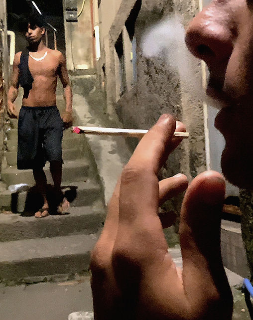Frame horizontal de homens em uma viela. Rosto de perfil aparecendo do nariz para baixo, próximo aos lábios entreabertos uma mão segurando um baseado entre os dedos. Ao fundo um jovem negro de corrente prateada, com a camisa pendurada no ombro, bermuda e chinelo desce a escadaria estreita entre casas de alvenaria a mostra.
