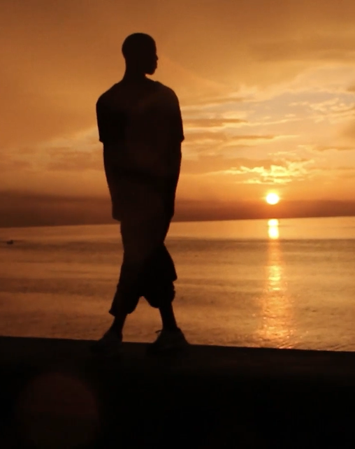 Frame horizontal de um homem na beira do mar no Sol poente. Sob uma luz alaranjado do fim do dia, silhueta de um homem alto de cabeça raspada, usa camiseta, calça e tênis, está em pé e olha para a direita com o queixo alto, está de pé com as pernas cruzadas uma em frente à outra. Atrás dele o brilho do mar calmo. Ao fundo à esquerda, silhuetas pequenas de prédios longínquos. O céu grande com esparsas nuvens e o sol arredondado quase no limiar.