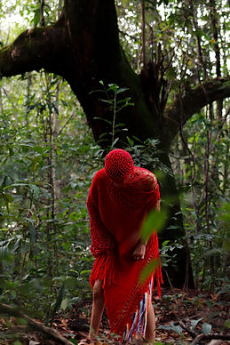 Frame do vídeo Desta Terra Viverei. Em meio à mata fechada uma figura humana revestida da cabeça aos joelhos por um manto de crochê vermelho. Tem o tronco e a cabeça inclinados para frente.
