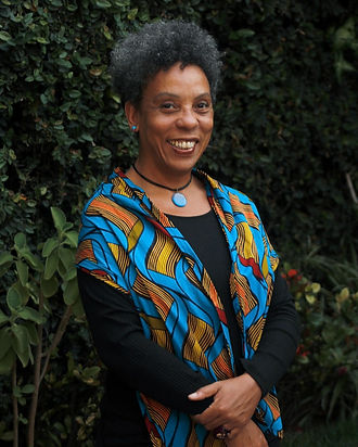Fotografia de Lilian da cintura para cima. Mulher cis negra de escuros cabelos crespos usando um quimono de capulana azul com detalhes em amarelo, laranja e vermelho. Sorri para a câmera.