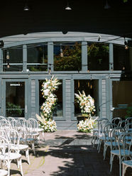 An outdoor wedding ceremony setup at Everett West in Portland featuring white chairs arranged on a patio beneath string lights, with two cascading floral installations of white orchids, baby’s breath, roses, and soft pastel blooms framing the ceremony space.