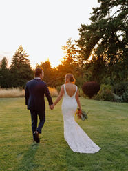 Bride and groom walk hand in hand across a large green lawn, heading toward a forest of evergreen trees, all framed by the golden hour sunlight.