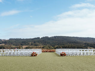 A ceremony set up with white chairs and red, orange, and peach florals. The backdrop is a stunning view of a vineyard in Oregon's wine country.