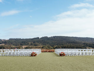 A ceremony set up with white chairs and red, orange, and peach florals. The backdrop is a stunning view of a vineyard in Oregon's wine country.