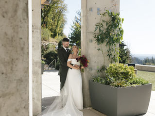 Bride in a flowy white dress and groom in a dark green suit looking into each others eyes smiling with concrete pillars and lots of greenery to frame the scene.