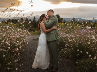 The couple shares a romantic kiss on a gravel path lined with delicate wildflowers, framed by a golden sunset and the rolling hills of Hood River in the background.