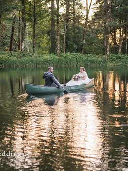 A bride and groom paddle around a lake in a canoe, surrounded by a forest with the evening sun peaking through the trees.