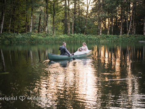 A bride and groom paddle around a lake in a canoe, surrounded by a forest with the evening sun peaking through the trees.