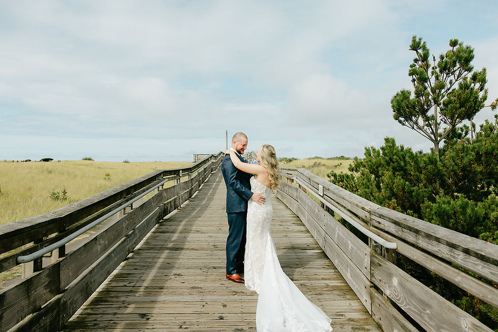 Bride and groom embracing each other on a beach boardwalk surrounded by greenery with the ocean as a backdrop.