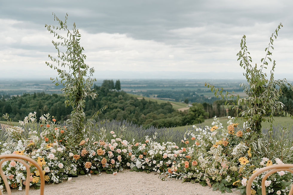 Ground floral arrangements in soft pink, peach, white, and yellow tones frame a vineyard ceremony aisle, with lush greenery and rolling hills stretching into the distance under a cloudy summer sky.