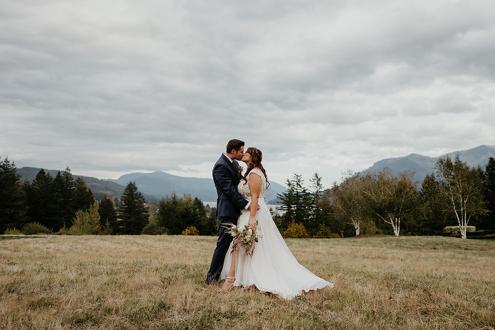 A bride and groom kissing in the middle of a field with trees, mountains, and the Columbia River as a backdrop.