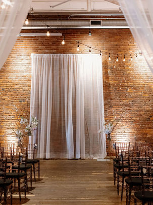 An indoor wedding ceremony setup at AXIS Pioneer Square with exposed brick walls, soft white draping framing the aisle, rows of dark wooden chairs, and warm string lights glowing overhead.