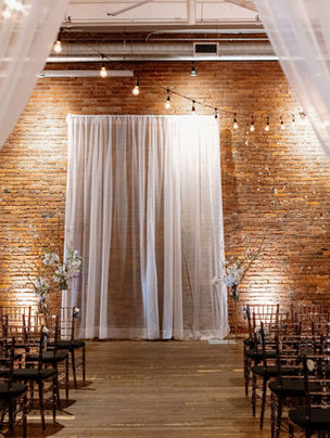 An indoor wedding ceremony setup at AXIS Pioneer Square with exposed brick walls, soft white draping framing the aisle, rows of dark wooden chairs, and warm string lights glowing overhead.
