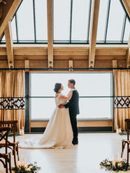 Bride in groom stand in front of a large window inside a lodge. Snow is falling in the background as they share a loving look.